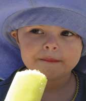 Photo of a three year old girl in a wide brimmed hat, eating an ice block. 