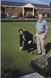 Doug Woolerton kneels on a bowling green watched by an elderly man. 
